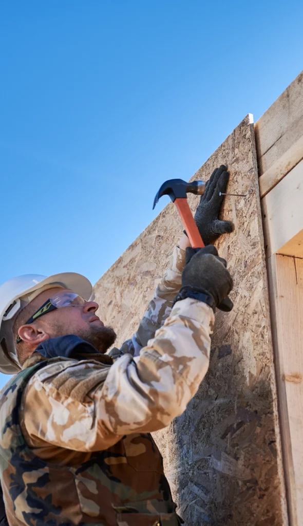 Man in camo work-wear using a hammer to secure a piece of OBS sheathing